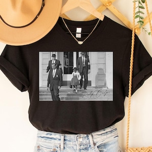 Op de afbeelding: Zwart T-shirt met een zwart-wit afbeelding van mensen die een trap aflopen. Het shirt wordt gecombineerd met een spijkerbroek, een beige hoed en een geweven crossbodytas. Een gouden ketting is zichtbaar.