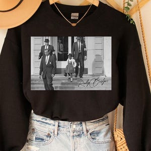 May include: Black crewneck sweatshirt featuring a black and white image of Ruby Bridges walking with U.S. Marshals. The image is signed "Ruby Bridges" in cursive. The sweatshirt is paired with denim shorts and a tan hat.