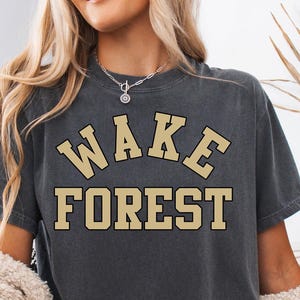 May include: Dark grey t-shirt with the text "WAKE FOREST" in a gold collegiate font. The shirt is worn with denim shorts and a cream-colored sweater. The overall theme is casual collegiate apparel.