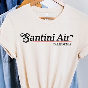 Puede incluir: Camiseta color crema con el texto "Santini Air CALIFORNIA" en negro y rojo. La camiseta está colgada en una percha de madera, con una camisa vaquera y un sombrero marrón en el fondo.
