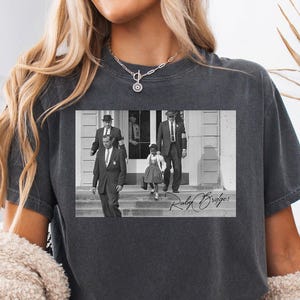 Könnte beinhalten: Dunkelgraues T-Shirt mit einem Schwarz-Weiß-Bild von Ruby Bridges, die von US-Marshals eskortiert wird. Das Bild enthält den Text "Ruby Bridges" in einer Schriftart. Das T-Shirt wird von einer Person getragen.