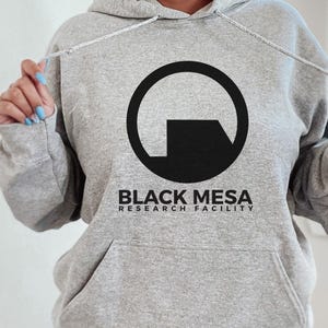 Peut inclure: Un sweat à capuche gris clair avec un motif circulaire noir et l'inscription "BLACK MESA RESEARCH FACILITY" en dessous. Le sweat à capuche est doté d'un cordon de serrage et d'une poche avant.
