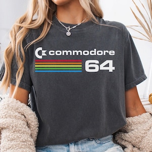 May include: Dark grey t-shirt featuring the Commodore 64 logo in white with a rainbow stripe design. The shirt is a vintage-style tee, perfect for retro gaming enthusiasts. The shirt is a comfortable, casual wear.