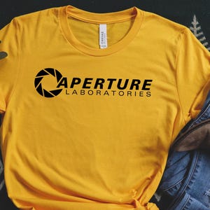 May include: Mustard yellow t-shirt with the Aperture Laboratories logo in black. The shirt is paired with blue jeans and brown ankle boots, arranged on a patterned surface.