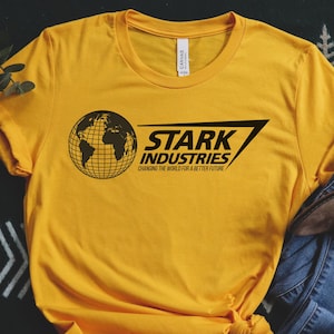 May include: Mustard yellow t-shirt with the text "STARK INDUSTRIES" and a globe graphic. The shirt is paired with blue jeans and brown boots, arranged on a patterned surface. The text on the shirt reads "Changing the world for a better future."