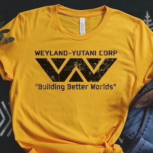 Könnte beinhalten: Senfgelbes T-Shirt mit dem Text "WEYLAND-YUTANI CORP" und dem Satz "Building Better Worlds" in Schwarz. Das Shirt wird mit einer blauen Jeans und braunen Stiefeletten kombiniert. Das Shirt hat einen Rundhalsausschnitt und kurze Ärmel.