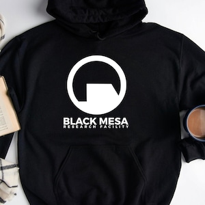 May include: Black hooded sweatshirt with a white graphic of a circle and a square, with the text "BLACK MESA RESEARCH FACILITY". A book, glasses, a plaid scarf, and a mug of coffee are also in the image.