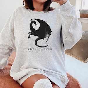 May include: A light gray sweatshirt featuring a black dragon graphic and the text "IT'S BEEN MY HONOR". The long-sleeved top is made of a soft material.
