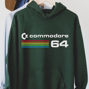May include: A dark green hoodie with the text "Commodore 64" in white, above a horizontal rainbow stripe. The hoodie has a front pocket and a hood. A brown hat and denim shirt hang on the rack behind the hoodie.