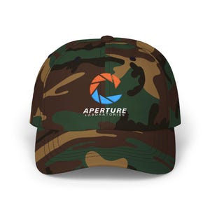 Puede incluir: Gorra de béisbol de camuflaje con el logotipo de Aperture Laboratories en naranja, azul y blanco. La gorra tiene una visera curva y un diseño clásico de seis paneles. El sombrero está hecho de una tela duradera.