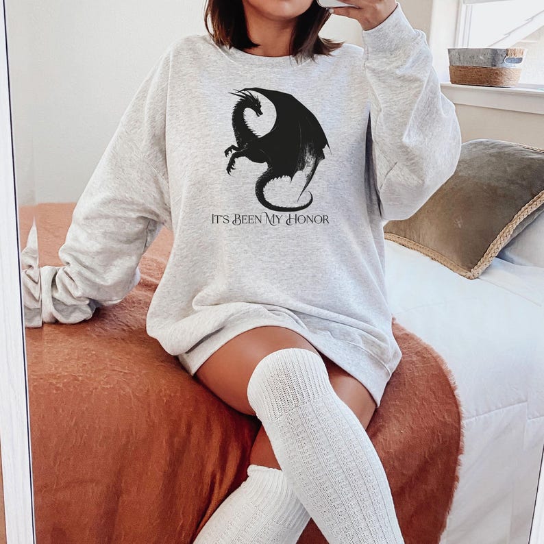 May include: A light gray sweatshirt featuring a black dragon graphic and the text "IT'S BEEN MY HONOR". The long-sleeved top is made of a soft material.