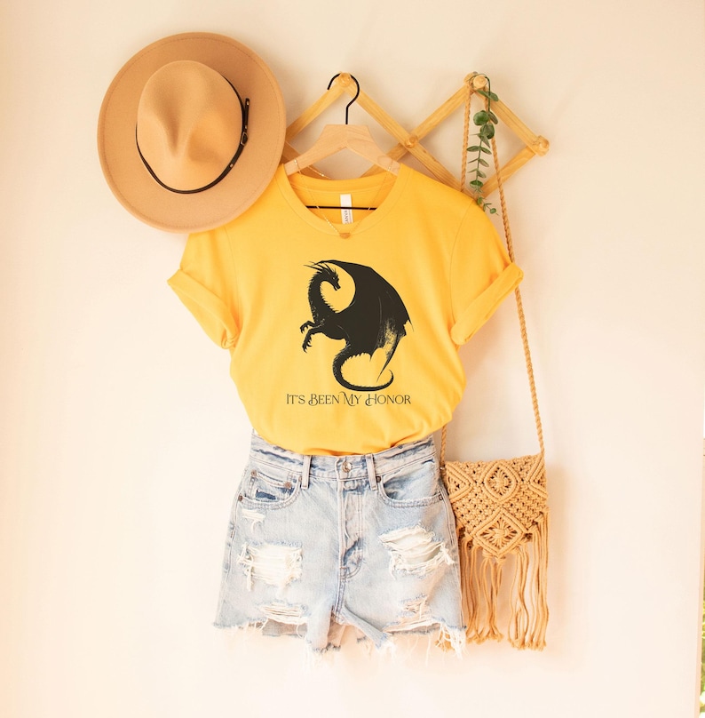 May include: A yellow v-neck t-shirt with a black dragon graphic and the text "IT'S BEEN MY HONOR." The shirt is paired with denim shorts, a tan hat, and a woven crossbody bag. A wooden hanger and decorative wall piece are also visible.
