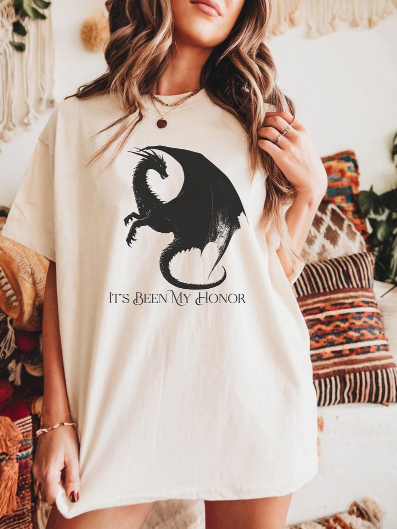 K&ouml;nnte beinhalten: Cremefarbenes T-Shirt mit einem schwarzen Drachenmotiv. Der Drache hat gro&szlig;e Fl&uuml;gel und einen geschwungenen K&ouml;rper. Unter dem Drachen steht der Text "IT'S BEEN MY HONOR". Das Shirt hat eine lockere Passform.