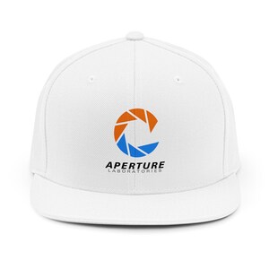 Puede incluir: Gorra de béisbol blanca de ala plana con el logotipo de Aperture Laboratories. El logotipo es un diseño estilizado en naranja y azul con el texto "APERTURE LABORATORIES" debajo. Un estilo clásico y casual.