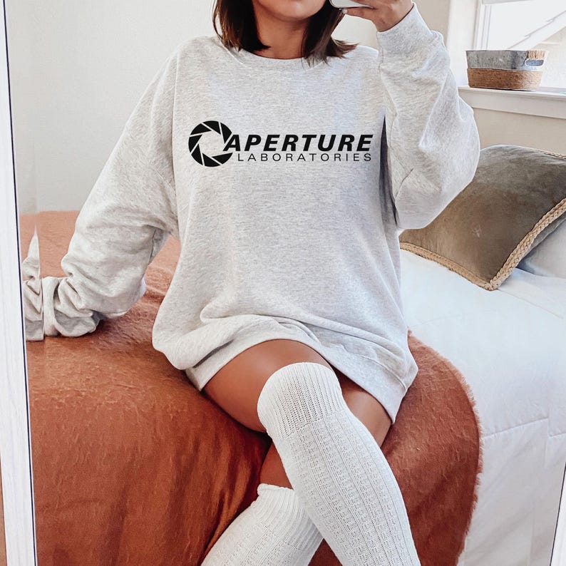 May include: A light gray sweatshirt with the text "APERTURE LABORATORIES" in black. The sweatshirt has long sleeves and a crew neck. The image shows a person wearing the sweatshirt.