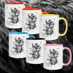 Pode incluir: Canecas de cerâmica brancas com uma ilustração a preto e branco de um guaxinim e um gambá a abraçar-se, com um fundo estrelado. Cada caneca tem um interior e uma pega de cor diferente: vermelho, laranja, amarelo, azul, preto e rosa.