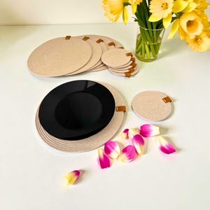 Pode incluir: Um conjunto de jogos americanos e porta-copos bege tecidos com detalhes em couro castanho. Um prato preto repousa sobre um jogo americano. Narcisos amarelos num vaso de vidro e pétalas de flores rosa e brancas completam a cena.