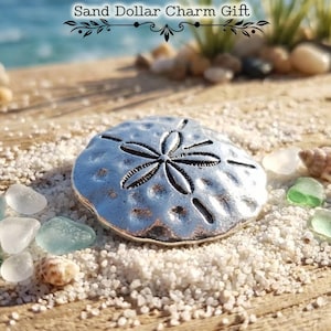 May include: A silver sand dollar charm with a detailed design, on white sand. The charm is surrounded by seashells and sea glass. The text "Sand Dollar Charm Gift" is above the charm.