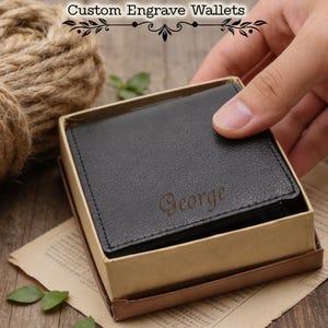 May include: Black leather wallet with the name "George" engraved on the front, presented in a cardboard gift box. The image also includes the text "Custom Engrave Wallets".