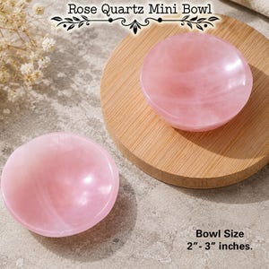 May include: Two smooth, polished rose quartz mini bowls with a light pink, translucent appearance. One bowl rests on a circular wooden base. The text Rose Quartz Mini Bowl is visible at the top, and Bowl Size 2-3 inches is printed below.