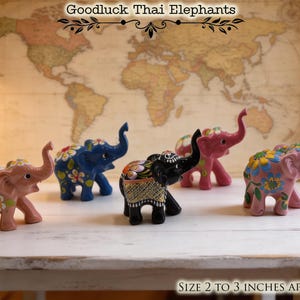 May include: A collection of ceramic elephant figurines, each approximately 2 to 3 inches in size. The elephants are painted in various colors, including pink, blue, and black, and decorated with floral patterns. The text "Goodluck Thai Elephants" is visible.