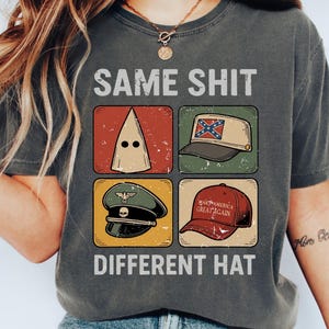 May include: Dark grey t-shirt featuring the text "SAME SHIT DIFFERENT HAT" above and below four images of hats. The images include a KKK hood, a Confederate flag hat, a military hat, and a "Make America Great Again" hat.