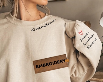 May include: A beige sweatshirt with the word "Grandma" embroidered in black script. The sleeve has names "James, Amelia, Emma" and a heart. A brown leather patch with "EMBROIDERY" is sewn on the front.