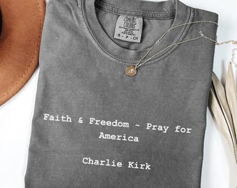 May include: A gray Comfort Colors t-shirt with the text "Faith & Freedom - Pray for America - Charlie Kirk" in white. A gold pendant necklace and a brown hat are also visible.