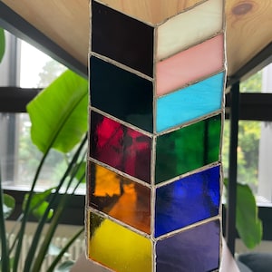 Könnte beinhalten: Ein sonnenfänger in Chevron-Form aus Buntglas mit einer Vielzahl von Farben, darunter Schwarz, Weiß, Rosa, Blau, Grün, Rot, Orange, Gelb und Lila. Das Glas ist mit silberfarbenem Metall umrahmt. Zum Aufhängen ist eine Kordel aus Naturfaser angebracht.