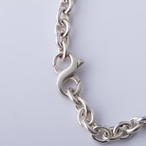 May include: Close-up of a silver chain necklace with a lobster clasp. The chain links are oval-shaped and connected. The clasp is in the shape of an infinity symbol. The necklace is on a white background.