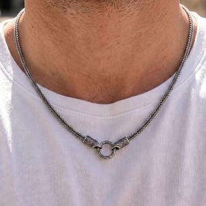 May include: A silver-toned wheat chain necklace featuring a textured, metallic finish. The chain connects at the front with two detailed, cylindrical end caps that secure a small, circular ring clasp, creating a cohesive and structured design.