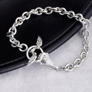 Op de afbeelding: Zilverkleurige kettingarmband met een unieke sluiting. De sluiting toont een skeletachtige hand die een ring vasthoudt, met vleugelaccenten. De armband is gemaakt van ovale schakels. De armband ligt op een zwarte ondergrond.