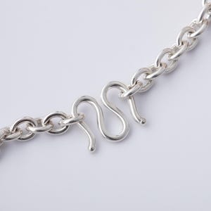 May include: Close-up of a silver chain necklace with a curved hook clasp. The chain links are oval-shaped and connected. The clasp is a stylized S-shape with a hook at the end. The necklace is on a white background.