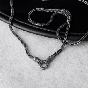 May include: A dark silver-toned chain necklace featuring a woven, textured link pattern. The necklace includes a circular spring ring clasp mechanism connected to two cylindrical end caps with etched, linear detailing along the surface.