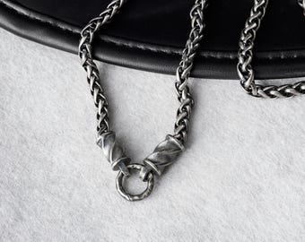 May include: A silver-toned wheat chain necklace featuring a dark, oxidized finish. The chain connects to two twisted, cylindrical metal end caps that secure a small, circular ring pendant at the center, creating a cohesive and metallic jewelry piece.