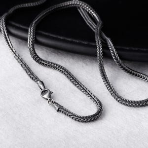 以下が含まれることがあります： 編み込みチェーンデザインのシルバーカラーのネックレス。ロブスタークラスプ留め。チェーンは暗く酸化した仕上げの金属製で、白い表面に表示されています。