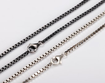 Op de afbeelding: Close-up van drie box chain kettingen in verschillende kleuren. Eén is zwart en de andere twee zijn zilver. Elke ketting heeft een karabijnsluiting. De kettingen worden diagonaal tegen een witte achtergrond weergegeven.