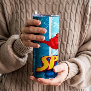 Pu&ograve; includere: Un bicchiere blu e bianco con un design da supereroe con la scritta "SP" in giallo e rosso. Il bicchiere ha una grafica a braccio rosso e una cannuccia bianca. Il bicchiere &egrave; tenuto da una persona che indossa un maglione beige.