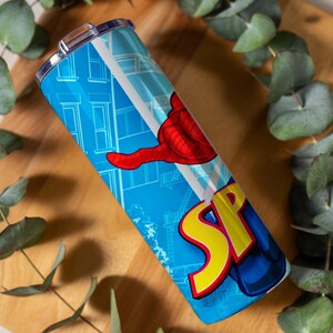 Pu&ograve; includere: Un bicchiere blu con un design a fumetti, con la parola "SP" in giallo e rosso e una mano rossa. Il design include uno sfondo di paesaggio urbano. Il bicchiere &egrave; in acciaio inossidabile e ha un coperchio argentato.