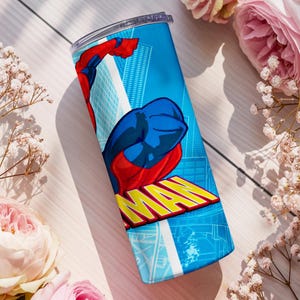 Pu&ograve; includere: Un bicchiere in acciaio inossidabile blu con un design da supereroe. Il design include una figura rossa e blu con la parola "MAN" in giallo. Lo sfondo presenta un paesaggio urbano. Il bicchiere &egrave; cilindrico e ha un coperchio trasparente.