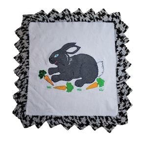May include: A white pillow with a black and white crocheted border. The pillow features an embroidered image of a black rabbit with blue eyes holding a carrot. The rabbit is surrounded by green leaves and orange carrots.