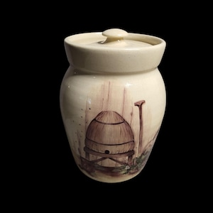 May include: Cream-colored ceramic jar with a lid. The jar features a hand-painted scene of a beehive, a wooden post, and a few flying bees. The jar has a rounded shape and a small knob on the lid. The background is a light beige color.