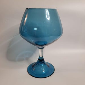 May include: A large, teal-colored glass goblet with a wide bowl and a clear stem. The goblet has a round base and a flared rim. The glass is smooth and reflects light, creating highlights. The overall design is elegant and modern.
