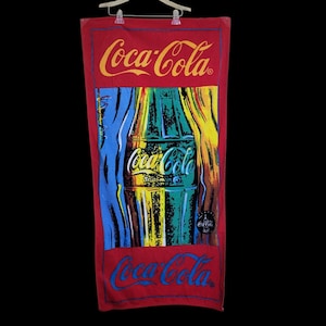 May include: Red Coca-Cola beach towel featuring a colourful graphic of a Coca-Cola bottle. The towel has the Coca-Cola logo in yellow at the top and bottom, with a vibrant bottle design in the centre. The design includes blue, green, and yellow.