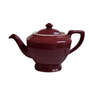 May include: A deep maroon ceramic teapot featuring a glossy finish, a matching lid with a small knob handle, a curved spout, and a sturdy side handle. The base of the teapot has a subtle fluted design, and the interior rim is a contrasting white.