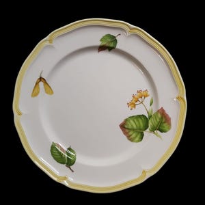 Può includere: Piatto in ceramica bianca con bordo smerlato e bordo giallo. Il piatto è decorato con elementi botanici dipinti a mano, tra cui foglie verdi, fiori gialli e baccelli. Il design è delicato ed elegante.