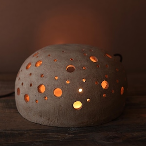 Puede incluir: Lámpara esférica de cerámica gris texturizada con numerosas aberturas circulares. La luz cálida emana del interior, creando un ambiente acogedor. La lámpara está sobre una superficie de madera, con un cable de alimentación negro.