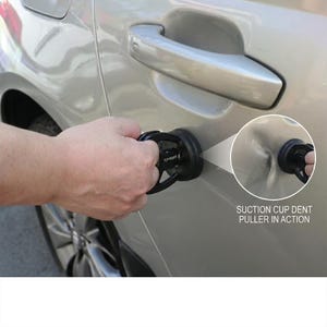 May include: A black suction cup dent puller is being used on a silver car door. The tool is designed to remove dents. The text "SUCTION CUP DENT PULLER IN ACTION" is visible in the image.
