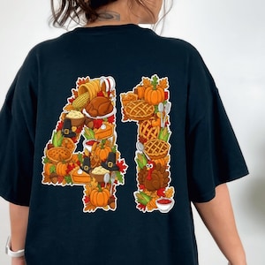 Może przedstawiać: Czarna koszulka z kolorowym motywem Święta Dziękczynienia. Numer "41" utworzony jest z ilustracji dyni, ciast, rogów obfitości i pieczonego indyka. Projekt jest obrysowany na biało.