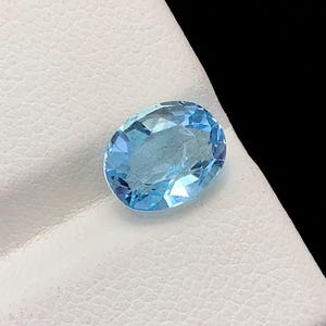 Puede incluir: Una gema ovalada de color azul claro con facetas, colocada sobre una superficie blanca texturizada. La gema tiene un aspecto brillante y claro, reflejando la luz. El fondo es negro, proporcionando contraste a la gema.